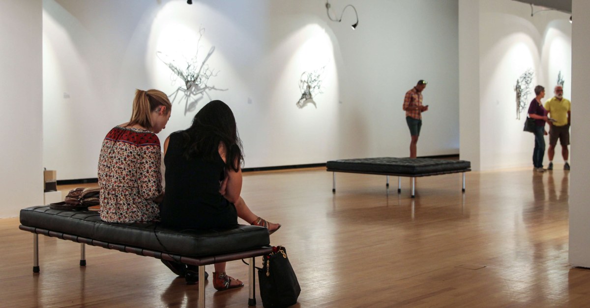 Two women in an all-white gallery, with three people in the background. 