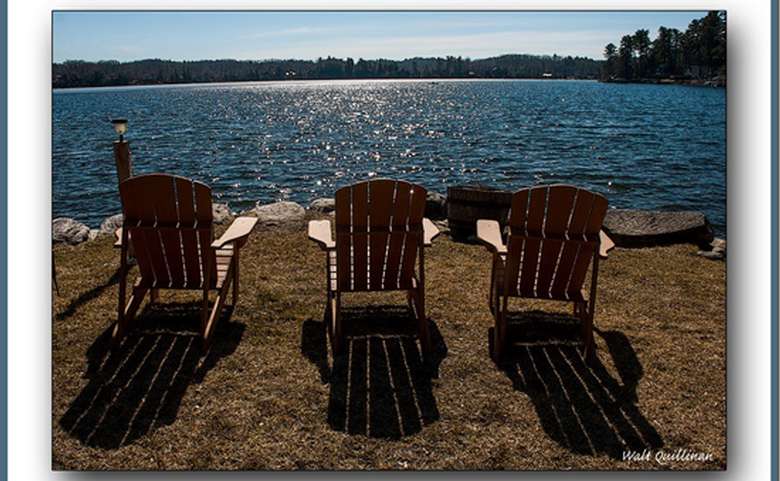 Three Adirondack chairs facing the lake. The sun is reflected off the water, creating a sparkling effect. There are trees and forests in the background.